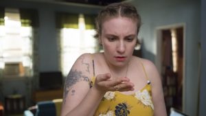 Woman in a yellow floral top looks down at her open hand with a puzzled expression; tattoo on her left upper arm is visible in a living room setting.