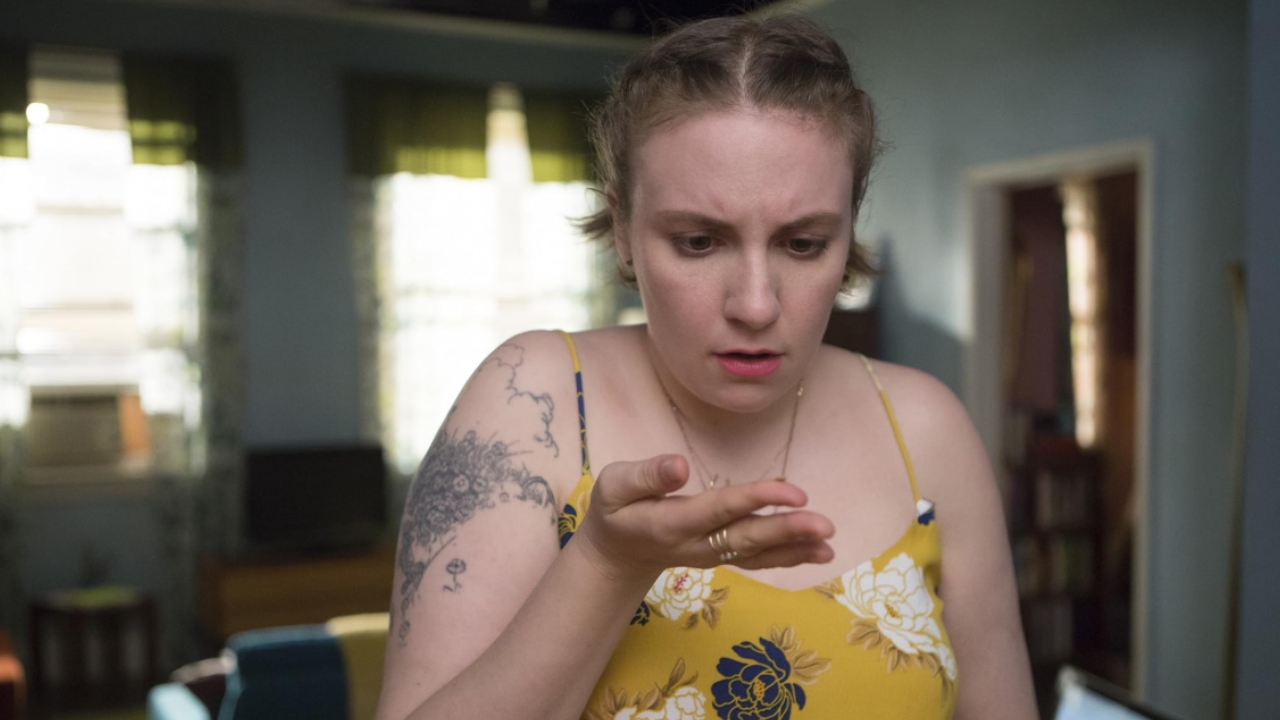 Woman in a yellow floral top looks down at her open hand with a puzzled expression; tattoo on her left upper arm is visible in a living room setting. 🎮 Outrun Gaming