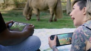 Two people sit on grass, play Steam Deck handhelds. Person on left turns to friend, smiles