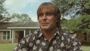 Man with blond hair wearing a paisley shirt sits outdoors in front of a house.